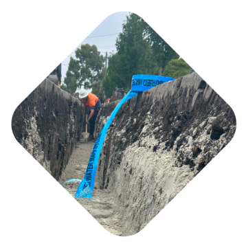 Worker in an orange vest digs a trench with a blue water pipe partially buried and visible. Trees and construction equipment are in the background.